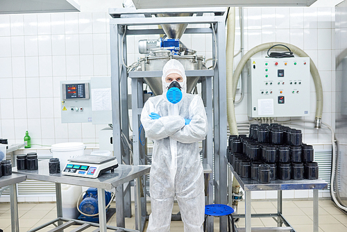 Full length portrait of confident factory worker wearing coverall and respirator  while standing against modern equipment with arms crossed, interior of manufacturing department