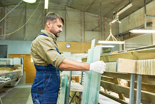 Side view portrait of mature man working in industrial workshop choosing material for cutting, wood and glass sheets, copy space