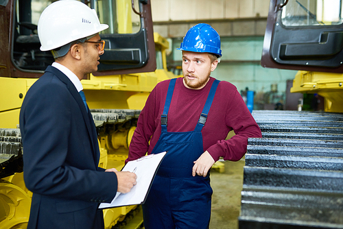 Handsome young worker wearing protective helmet and overall showing heavy equipment to mixed-race customer while standing at spacious warehouse of modern factory