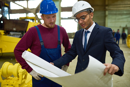 Concentrated factory worker and his mixed race superior studying blueprint and sharing ideas with each other while standing at spacious production department
