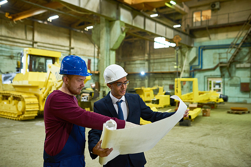 Young mixed race investor and bearded industrial engineer wearing hardhats gathered together at spacious production department of heavy equipment factory and studying blueprint