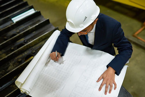 High angle view of concentrated young engineer wearing protective helmet and hardhat making necessary corrections in blueprint while standing at spacious production department of modern factory.