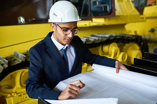 Waist-up portrait of handsome young businessman wearing suit and protective helmet studying blueprint with concentration while standing at warehouse of heavy vehicle factory