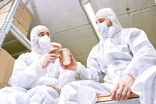 Low angle view of male warehouse workers wearing safety masks and coveralls clinking paper cups together while taking coffee break