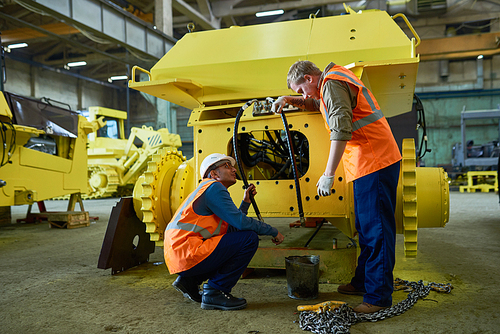 Two young technicians wearing reflective vests and overalls adjusting engine features of heavy vehicle while working at spacious production department of factory