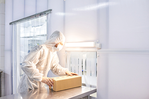 Male worker wearing coverall and safety mask standing at laboratory bench and packing pill bottles in cardboard box