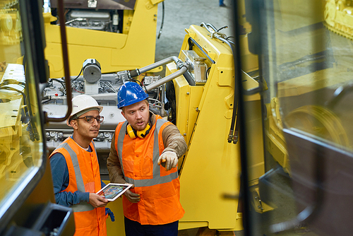 Bearded industrial engineers wearing protective helmets and reflective vests using digital tablet while discussing results of accomplished work, view from cabin of tractor