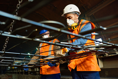 Low angle view of steel plant workers wearing reflective vests and hardhats lifting heavy sheet metal with help of crane, interior of production department on background