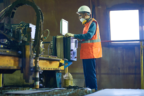 Profile view of concentrated young machine operator wearing hardhat and reflective vest wrapped up in work, interior of spacious production department on background
