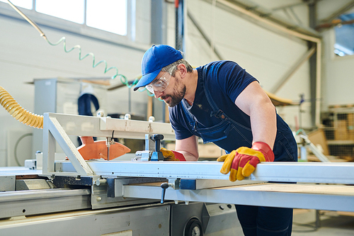 Serious concentrated young bearded worker of furniture factory wearing blue cap and safety goggles working with modern measurement equipment at plant