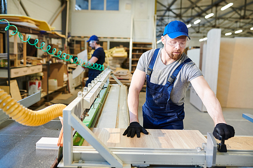 Serious busy young bearded man in blue cap and uniform standing at assembling machine and joining furniture piece while working at furniture factory