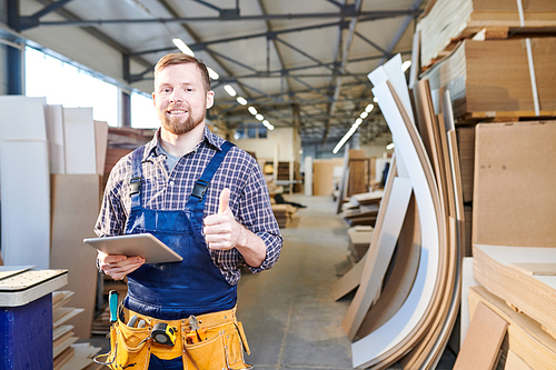 Happy excited young bearded repairman in overall wearing tool belt standing in warehouse of furniture details and using tablet while showing thumb-up and  in factory workshop