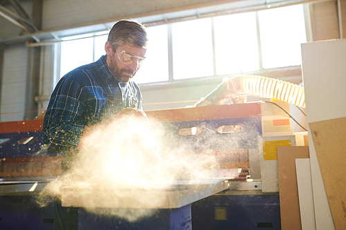 Serious busy handsome middle-aged bearded man in protective eyewear standing at workbench and cleaning wooden piece from sawdust while producing particleboard at plant