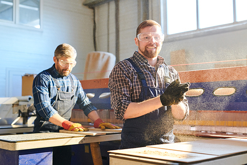 Cheerful excited handsome young bearded man in overall wearing work gloves clapping hands while removing sawdust, he working at furniture factory