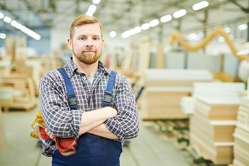 Smiling content confident handsome young bearded worker wearing overall standing at furniture factory and