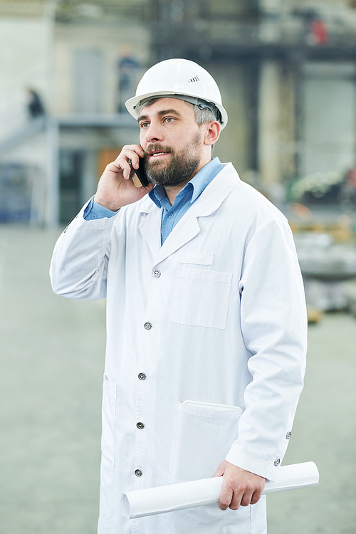 Portrait of mature bearded man wearing hardhat speaking by smartphone standing in industrial workshop