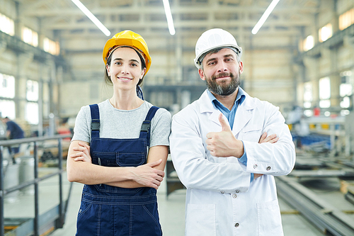 Waist up portrait of two smiling factory workers showing thumbs up and   while posing in industrial workshop