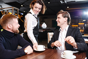 Smiling attractive young waitress with short hair  holding tray and putting cup on table while serving tea to businessmen