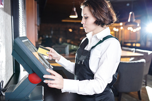Serious concentrated young waitress with short hair standing at restaurant desktop computer and using plastic card while choosing dish on POS terminal