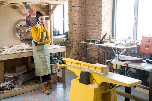 Full length portrait of modern young woman speaking by phone while working in loft-styled carpenters manufactory, copy space