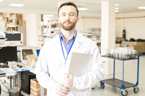 Waist-up portrait of handsome middle-aged engineer wearing lab coat holding laptop in hands and  while standing at spacious production department of manometer factory.
