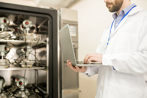 Close-up shot of concentrated lab technician standing at control cabinet, taking reading from pressure sensors and taking notes on laptop