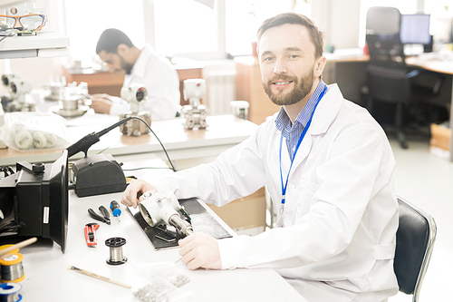 Cheerful confident bearded male repairing engineer in lab coat sitting at table with tools and dissembled measuring device and 