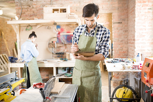 Portrait of focused modern carpenter holding notebook and sketching or writing in joinery shop, copy space