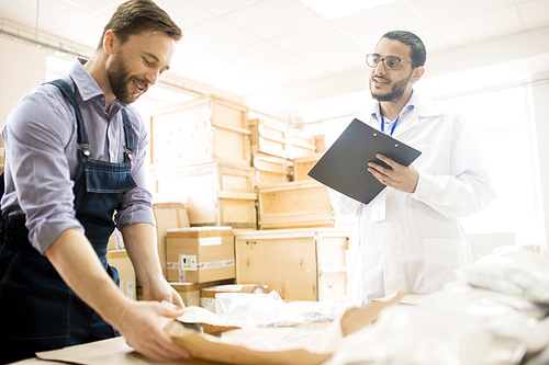 Handsome bearded warehouse worker packing detail while his superior wearing lab coat keeping eye on him and giving necessary recommendations