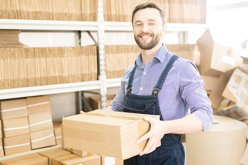 Portrait shot of middle-aged worker wearing checked shirt and denim jumpsuit holding cardboard box in hands and  with toothy smile, factory warehouse interior on background