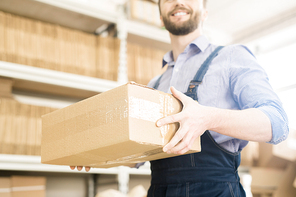 Close-up shot of bearded warehouse worker wearing checked shirt and denim jumpsuit  carrying cardboard box while preparing production lot for delivery