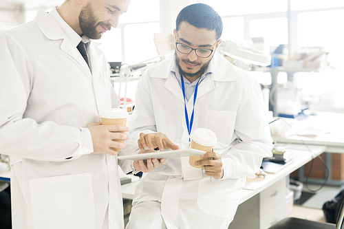 Serious concentrated handsome multiethnic engineers in lab coats drinking coffee while viewing data on tablet and discussing device production in factory office