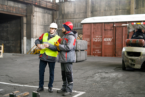 Full length portrait of two industrial workers wearing warm jackets and hardhats discussing production standing in workshop, copy space