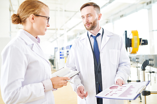 Bearded middle-aged dairy plant worker and his pretty colleague wearing white coats standing at modern production department while taking inventory