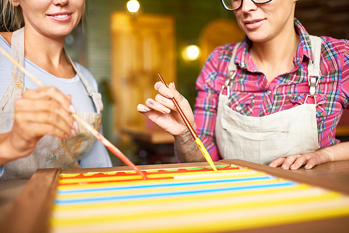 Mid-section  portrait of two young women  enjoying  working together in art studio, painting shutters for new art project, focus on female hands holding paintbrushes