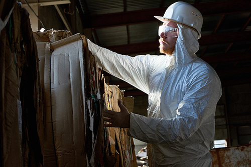 Side view portrait of factory worker wearing biohazard suit sorting reusable cardboard on waste processing, copy space