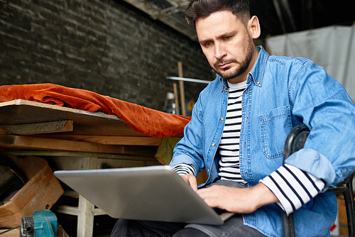 Portrait of handsome mature  man using laptop sitting in chair  in barn-like workshop, copy space