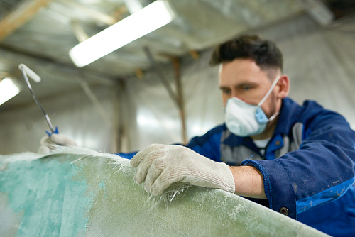 Portrait of mature man wearing protective mask repairing boat while working in yacht workshop, focus on foreground