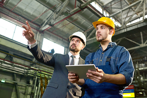 Waist up portrait of handsome businessman pointing up while discussing something with factory worker holding digital tablet, copy space