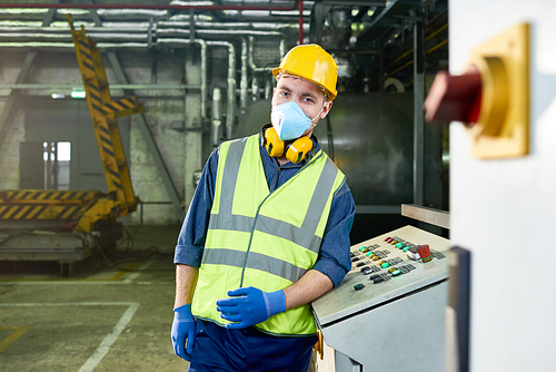 Waist up portrait of young factory worker wearing hardhat and protective mask  while posing in plant workshop standing by control panel, copy space