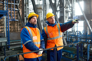 Group of bearded inspectors wearing uniform and protective helmets standing at production department of modern plant while making inspection, one of them pointing at something