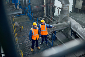 High angle view of bearded engineers wearing uniform and protective helmets standing at spacious production department of modern plant and brainstorming on joint work