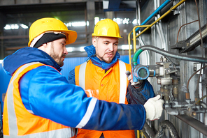 Group of bearded technicians wearing uniform and hardhats gathered together at spacious production department of modern plant and adjusting equipment