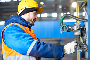 Profile view of highly professional technician wearing uniform and protective helmet adjusting equipment while standing at spacious production department of modern plant