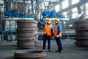 Bearded inspector wearing reflective vest and hardhat pointing at something while young technician giving tour of modern factory, interior of spacious production department on background