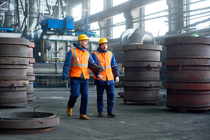 Team of hard-working technicians wearing hardhats and reflective vests walking along spacious production department of modern plant and discussing results of accomplished work