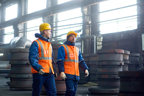 Brutal young machine operators wearing protective helmets and uniform chatting with each other while walking along production department of modern plant