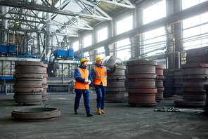 Bearded young worker wearing reflective vest and hardhat showing results of accomplished work to his superior while walking along production department of modern plant