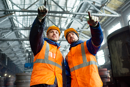 Group portrait of confident bearded workers wearing hardhats and reflective vests pointing at something with index fingers while standing at production department of modern plant