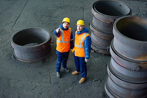 High angle view of bearded worker wearing reflective vest and hardhat pointing upwards while standing next to his colleague at production department of modern plant